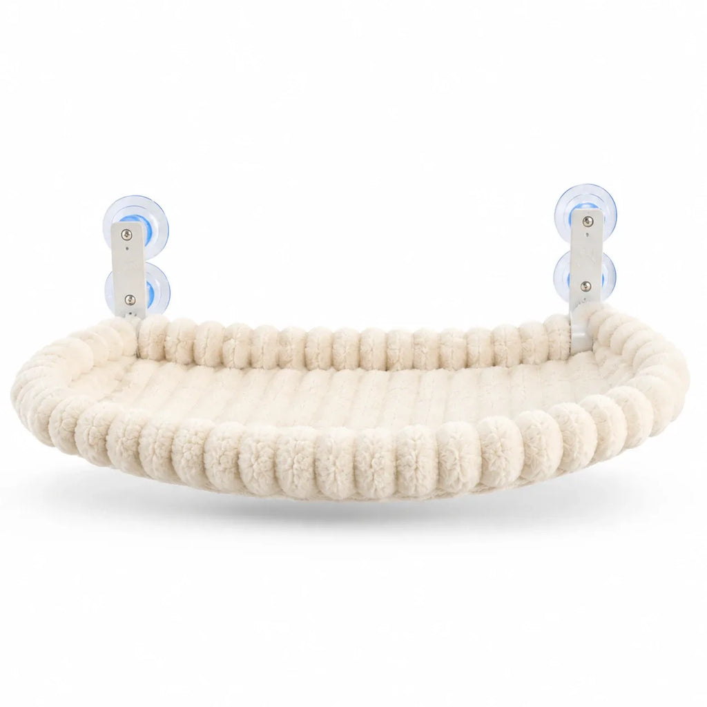 Cream plush cat window hammock with pom-pom border and paw-print suction cups, isolated three-quarter view